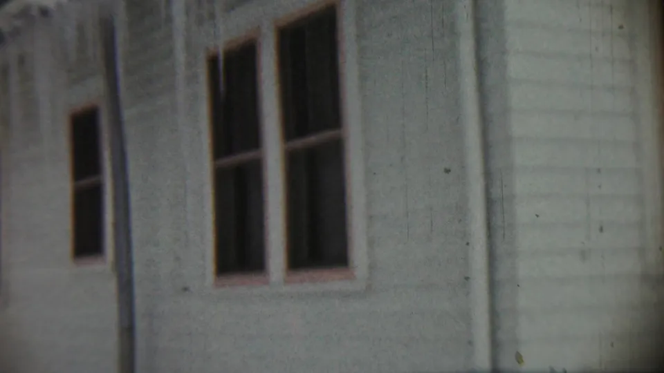 1959 home-movie frame showing dark window openings and light siding on a weathered residential exterior in Chadron, Nebraska.
