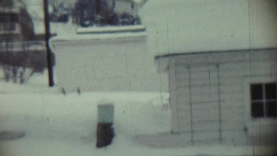 Chadron, Nebraska 1959 frame showing a snow-covered front lawn, bare trees, a small foreground post, and part of a house exterior.