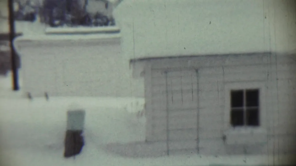 Late-1950s Chadron, Nebraska home-movie frame showing a house wall, dark window, bare tree trunk, and snow-covered lawn.