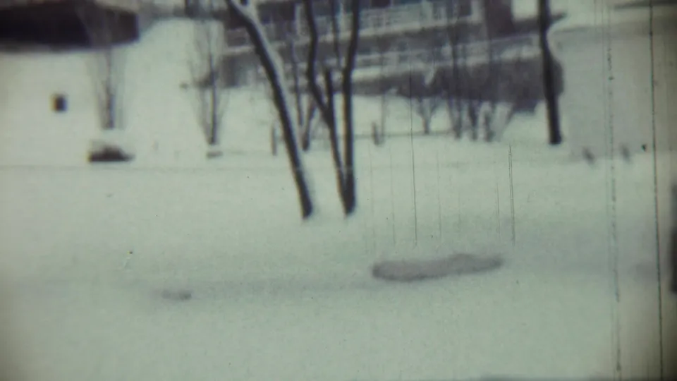 1959 home-movie frame from Chadron showing a snow-blanketed yard, clustered bare trees, and a pale structure in the background.