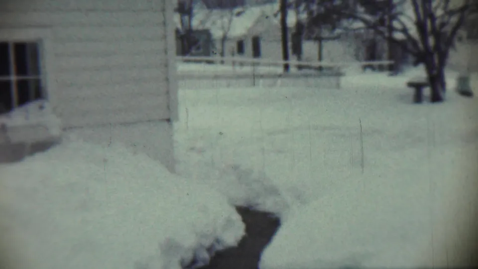1959 home-movie view of a Chadron house exterior, windows, deep snowbanks, fence line, and bare winter trees.