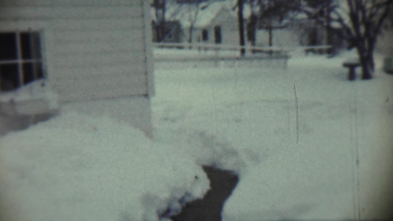 Snow-covered yard beside a house, with a dark icy channel cutting through deep drifts.
