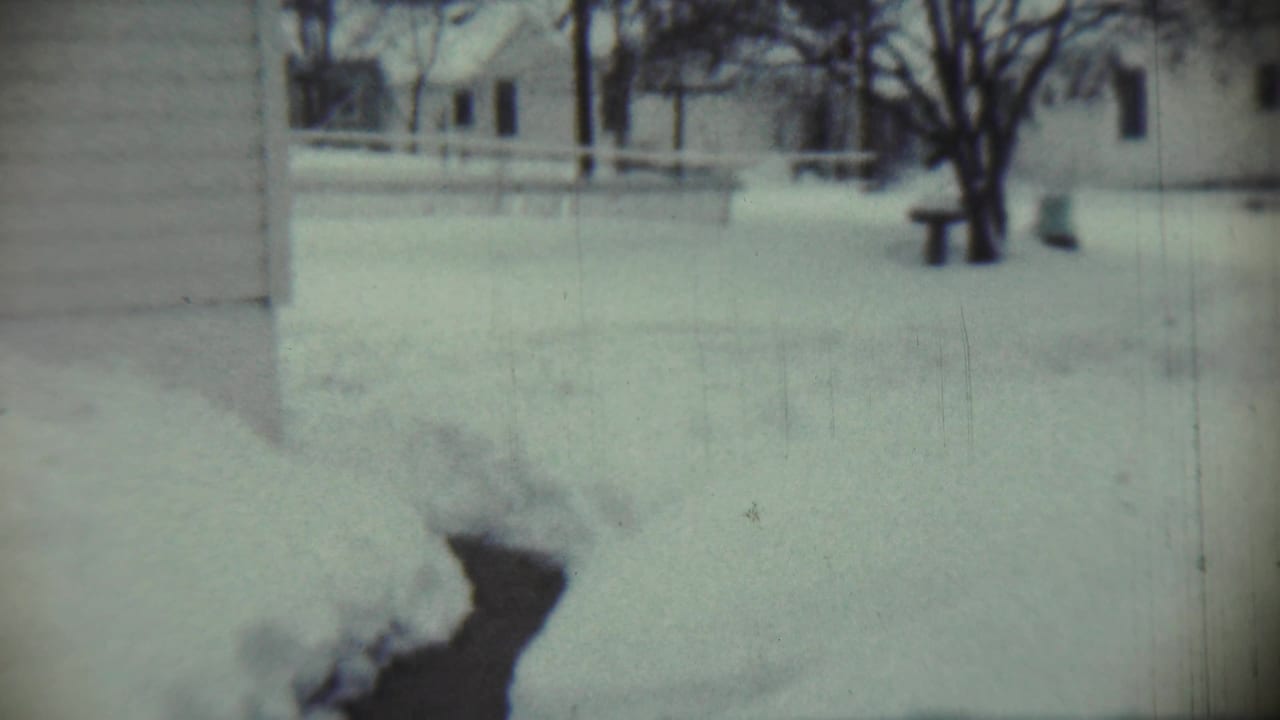 1959 Chadron, Nebraska winter frame with a snow-covered yard, bare trees, house walls, and a small bench or post in the distance.