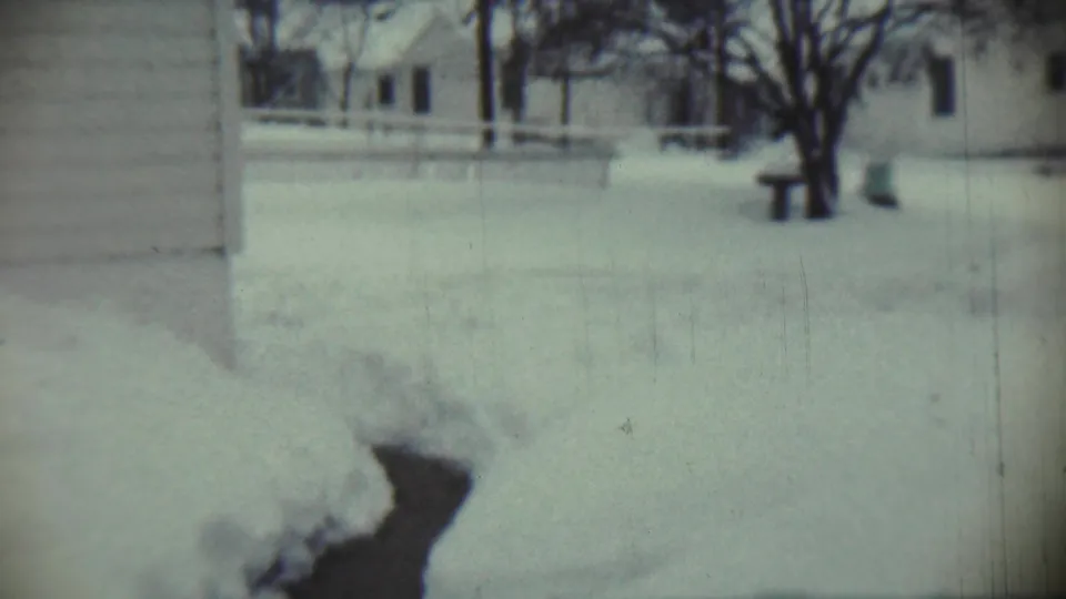 1959 Chadron, Nebraska winter frame with a snow-covered yard, bare trees, house walls, and a small bench or post in the distance.