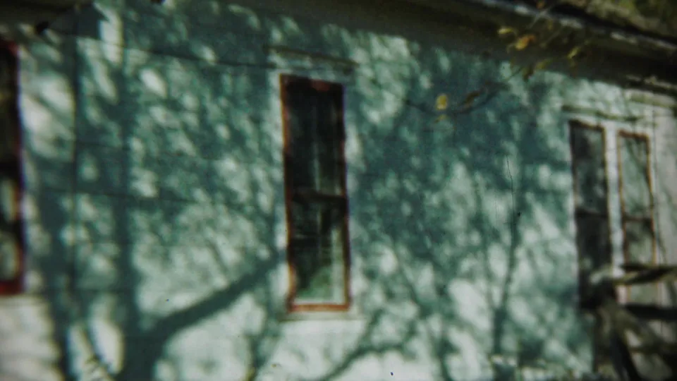 Chadron house exterior shows a tall red-trimmed window, door area, small porch rail, and winter branch shadows on the siding.