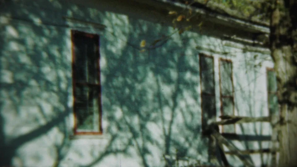 Weathered Chadron house exterior with narrow windows, porch steps or railing, and bare tree shadows across the pale siding.