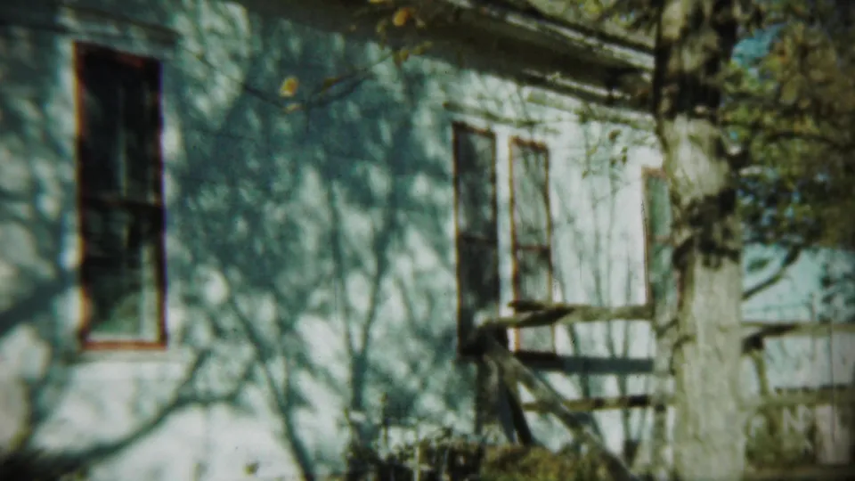 1959 Chadron, Nebraska 8mm frame of a white house exterior with a dark window, tree trunk, and sunlit shadows.