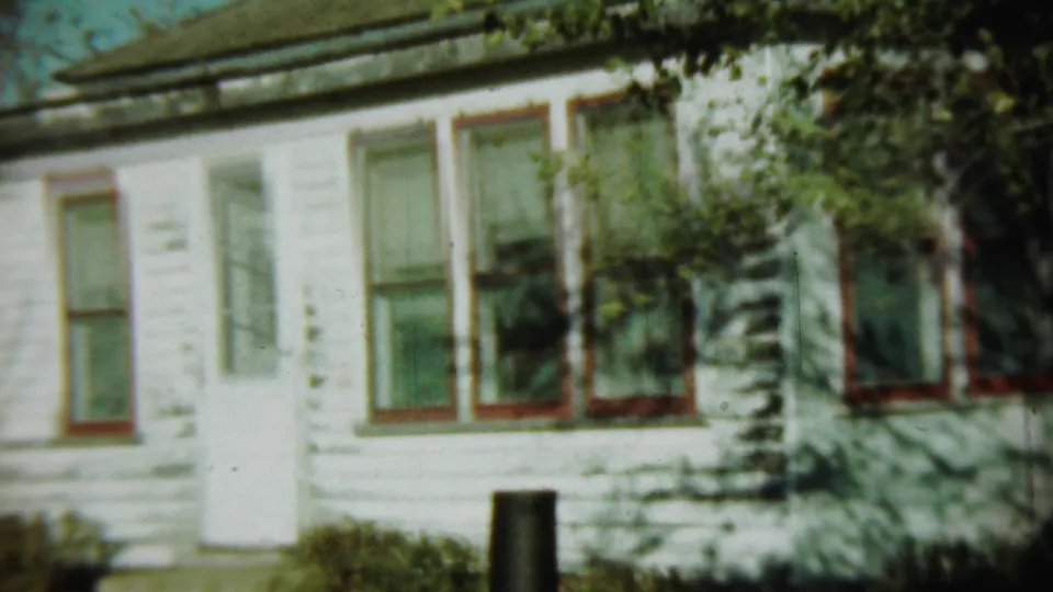 1959 Chadron, Nebraska 8mm frame showing a weathered white clapboard house with red-trimmed windows and leafy shadows.