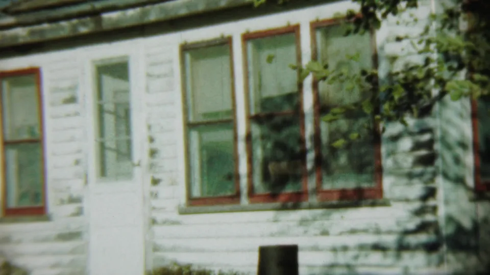 1959 Chadron, Nebraska home-movie frame showing a weathered white clapboard house with red-trimmed windows, a white door and leafy shadows.
