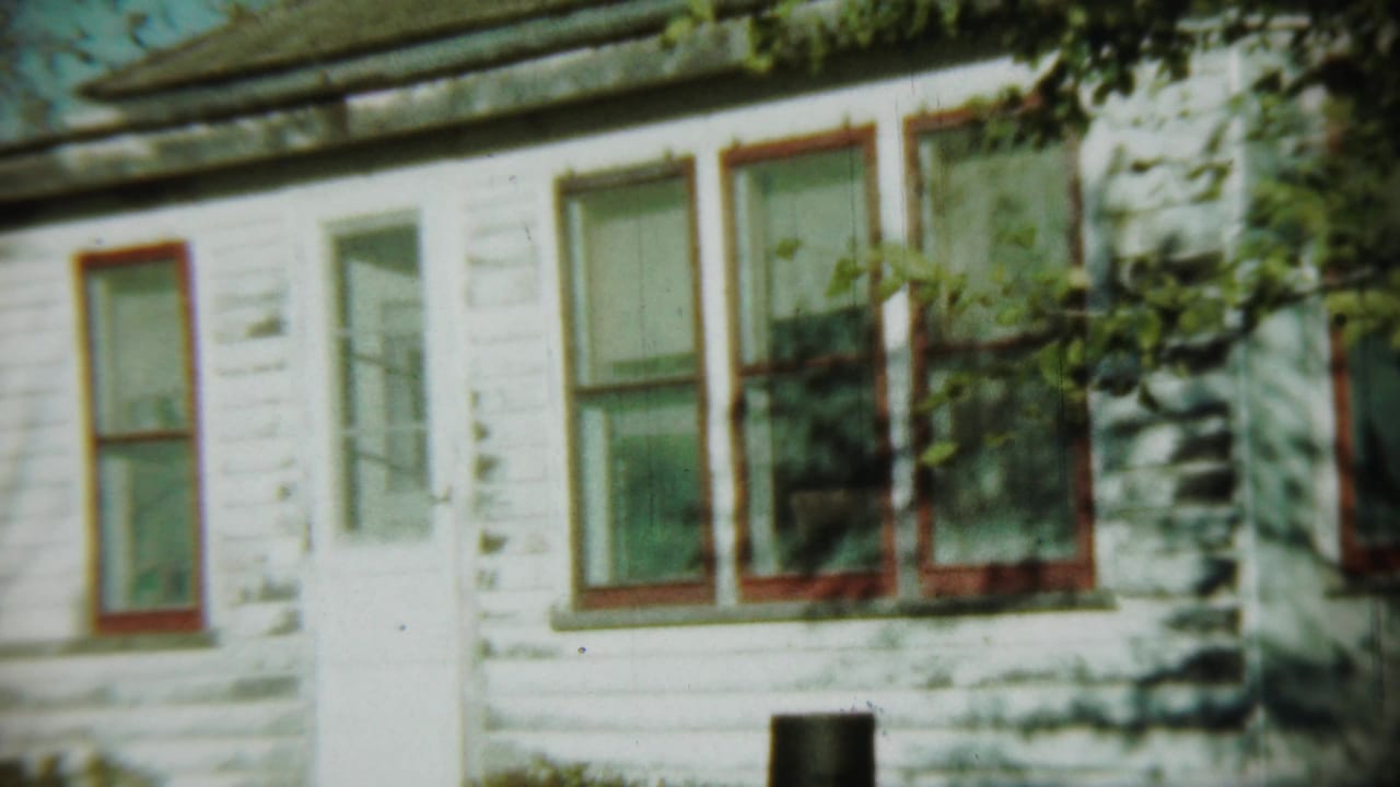 1959 Chadron, Nebraska frame of a modest white clapboard house with red window trim, a pale door and overhanging greenery.