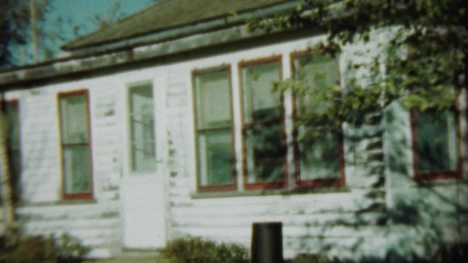 1959 Chadron, Nebraska frame showing a white clapboard residential exterior with red-trimmed windows, foliage and a dark post or barrel near the wall.