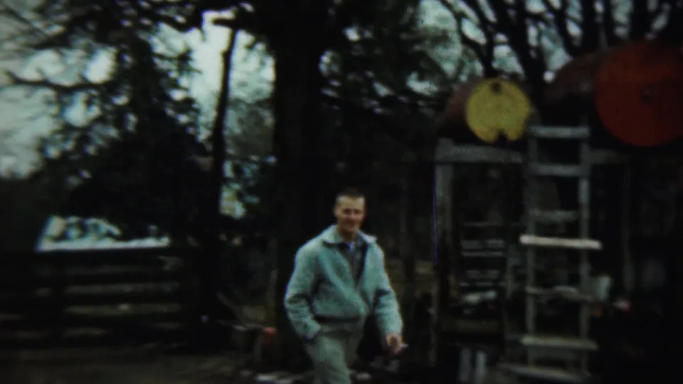 Adult man in a light jacket walks near a wooden structure with yellow and red circular objects in 1959 Chadron footage.