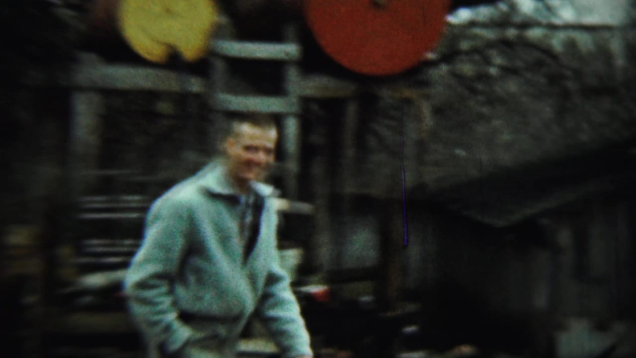 Man in a light jacket stands near barrels and a ladder in a rural yard.