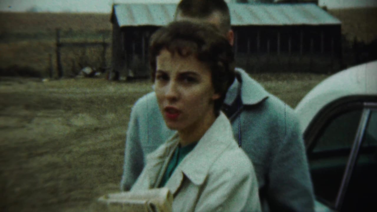 Woman in a white coat stands near a man and a white car in a farmyard.