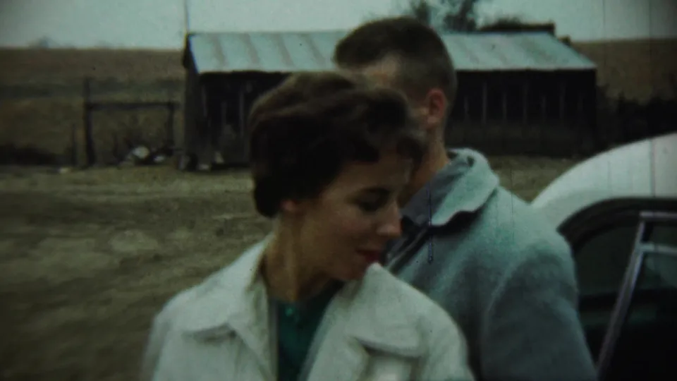 1959 Chadron, Nebraska home-movie frame of a woman looking down beside a man and a white car in a farmyard.
