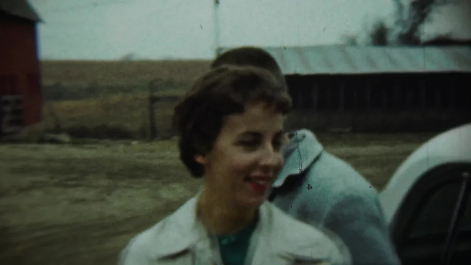 In 1959 Chadron, Nebraska footage, a young woman in a white coat smiles in a farmyard with cars, a red barn, and open fields behind her.