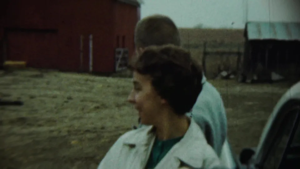 The woman turns away beside a man in a farmyard in Chadron, Nebraska, with a red barn, shed, fencing, and an older car visible.