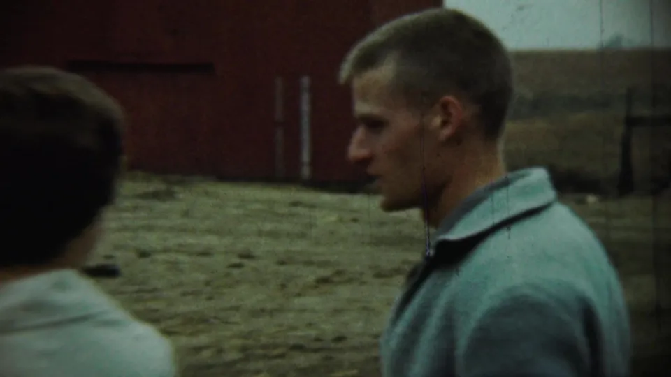 A man in a light jacket is seen in profile walking past a red barn and open farmyard as the woman in a white coat moves out of view.