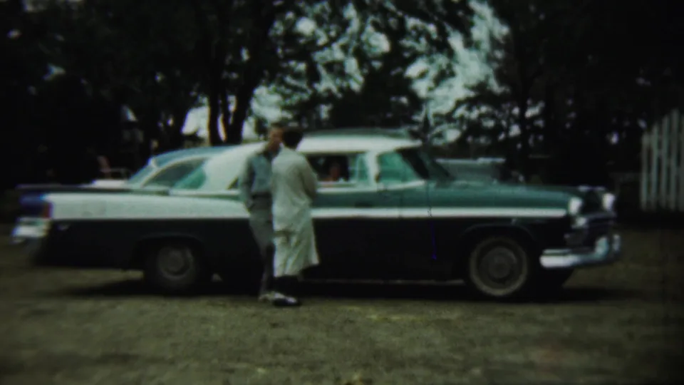 A 1959 Chadron, Nebraska home-movie frame shows a couple beside the passenger window of a green and white sedan in a rural yard.