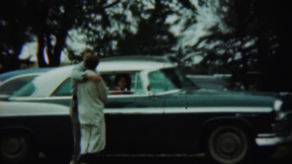 A closer 1959 Chadron, Nebraska frame shows the couple embracing or leaning together while speaking to someone inside the sedan.
