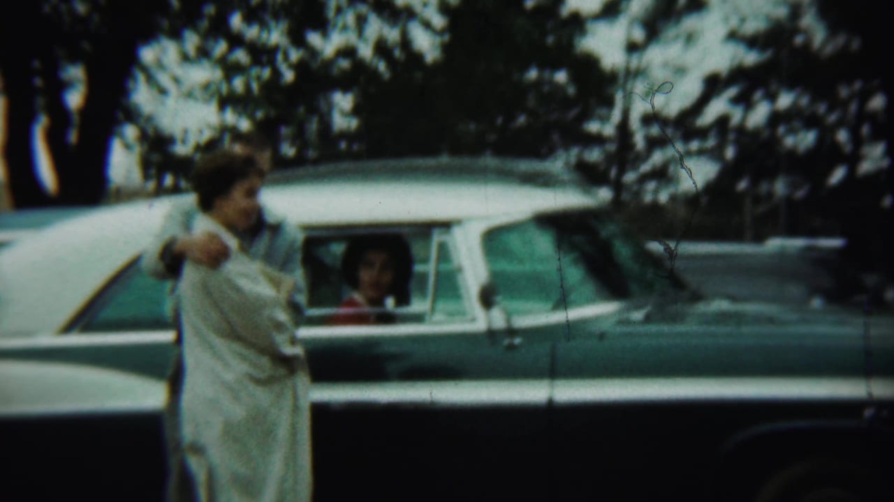 Adults stand beside a two-tone sedan while a passenger looks from the rear window.