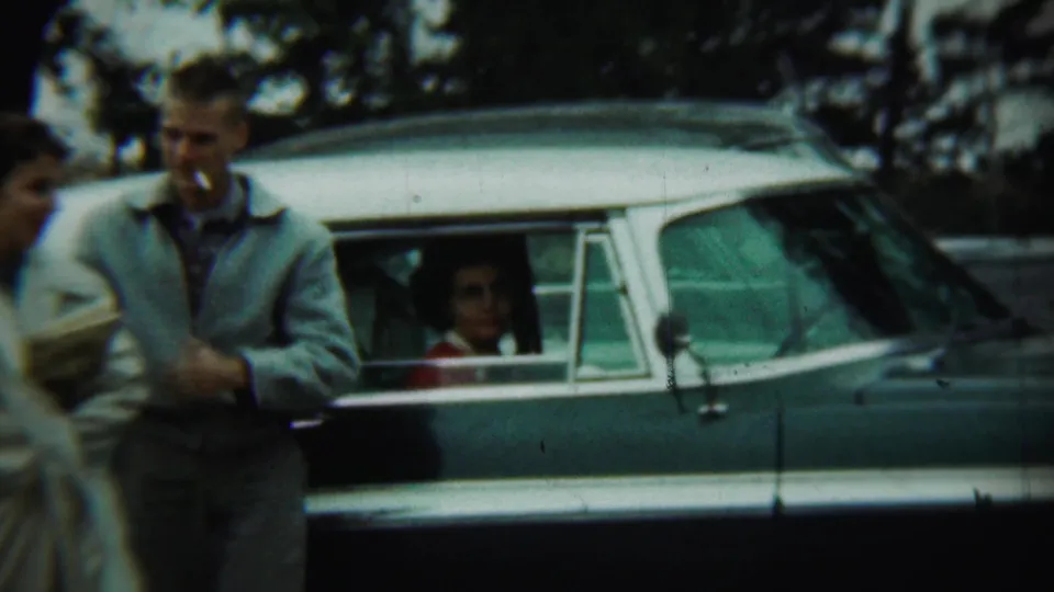 A man in a light jacket stands beside the mid-century sedan as another adult moves through the foreground in the 1959 Chadron footage.