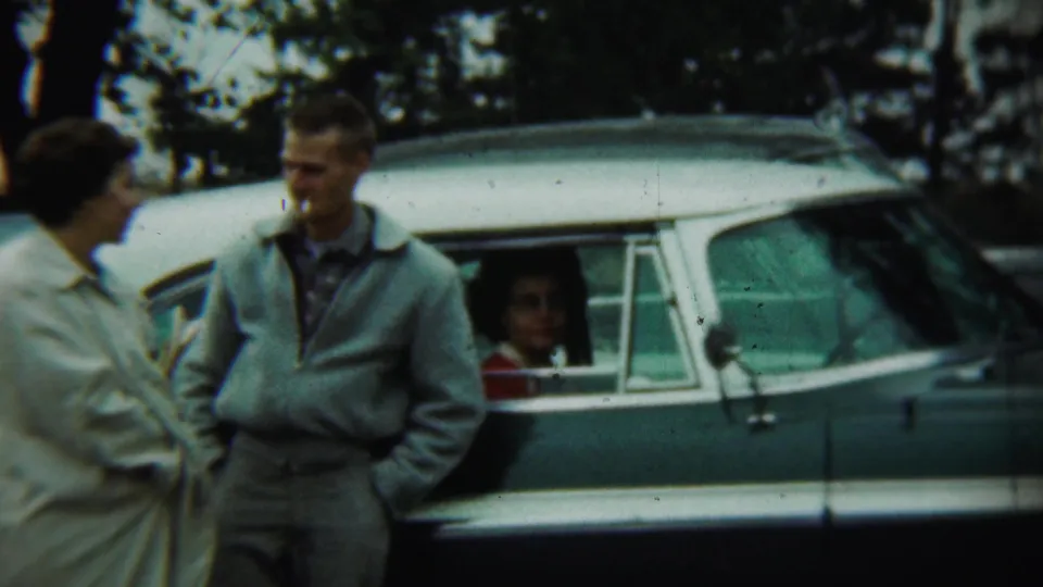 Two adults stand close together beside the two-tone sedan, with a rear-seat passenger visible through the window in the Chadron home movie.