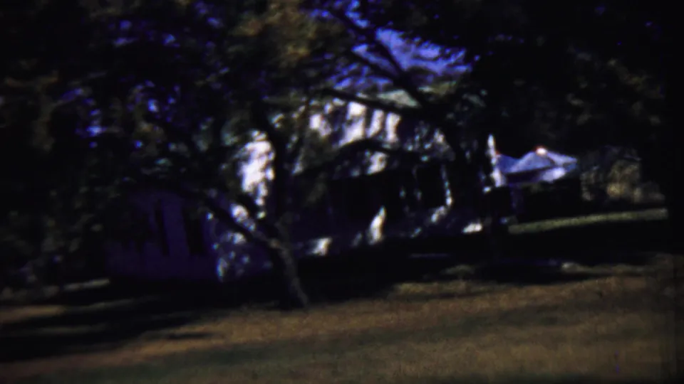Rural Kansas house partly obscured by mature trees, with a shaded yard and soft 8mm color shift.