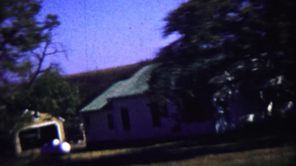1959 rural Kansas frame with a white building, green roof, trees, lawn, and the edge of a nearby vehicle or structure.