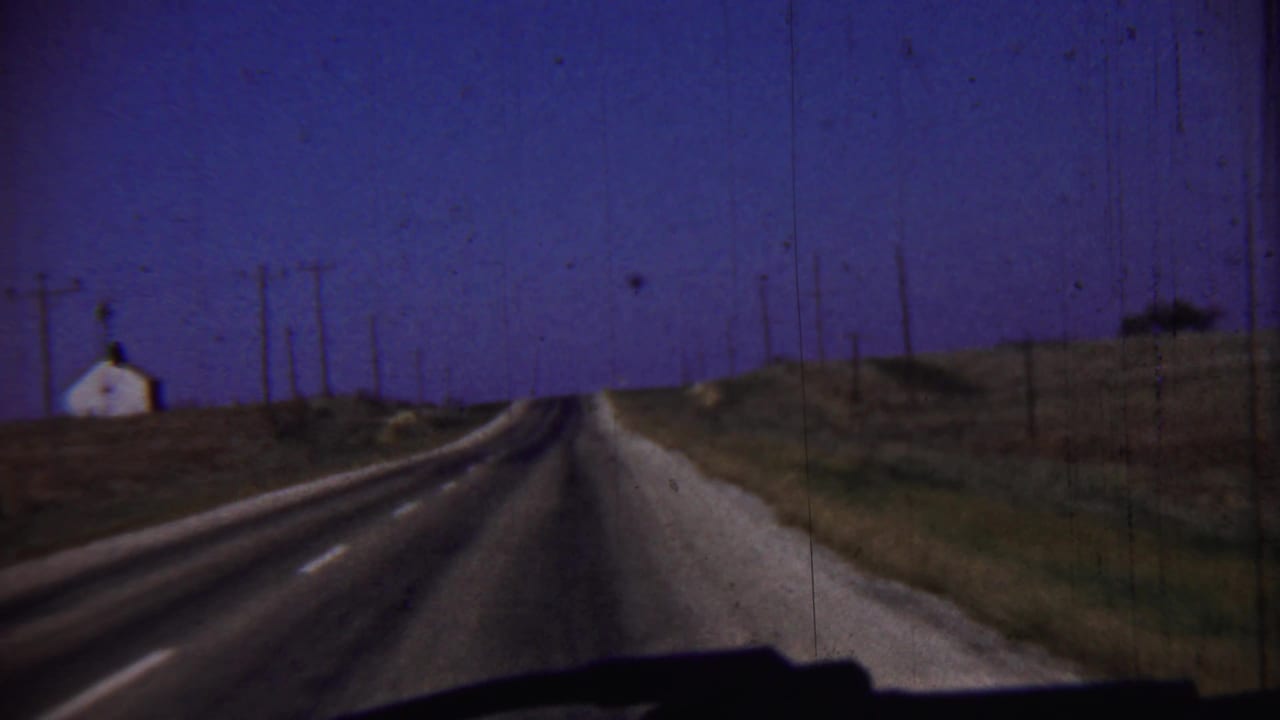 Two-lane rural road seen from a car, with a small white building and utility poles under a dark sky.