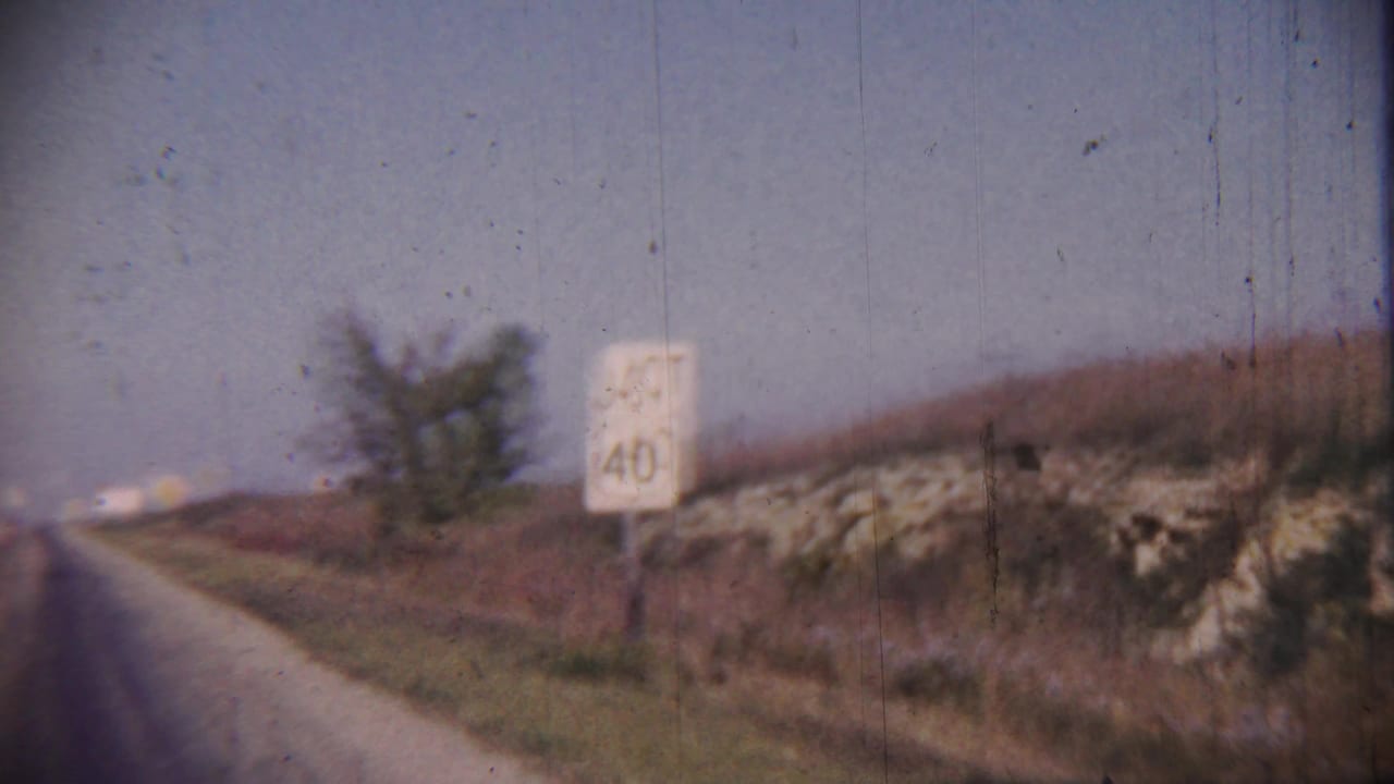 1959 Cheyenne County, Kansas road view with a white JCT 40 sign, grassy shoulder, low rocky bank, tree, and hazy sky.