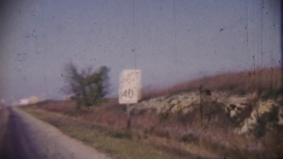 1959 Cheyenne County, Kansas road view with a white JCT 40 sign, grassy shoulder, low rocky bank, tree, and hazy sky.