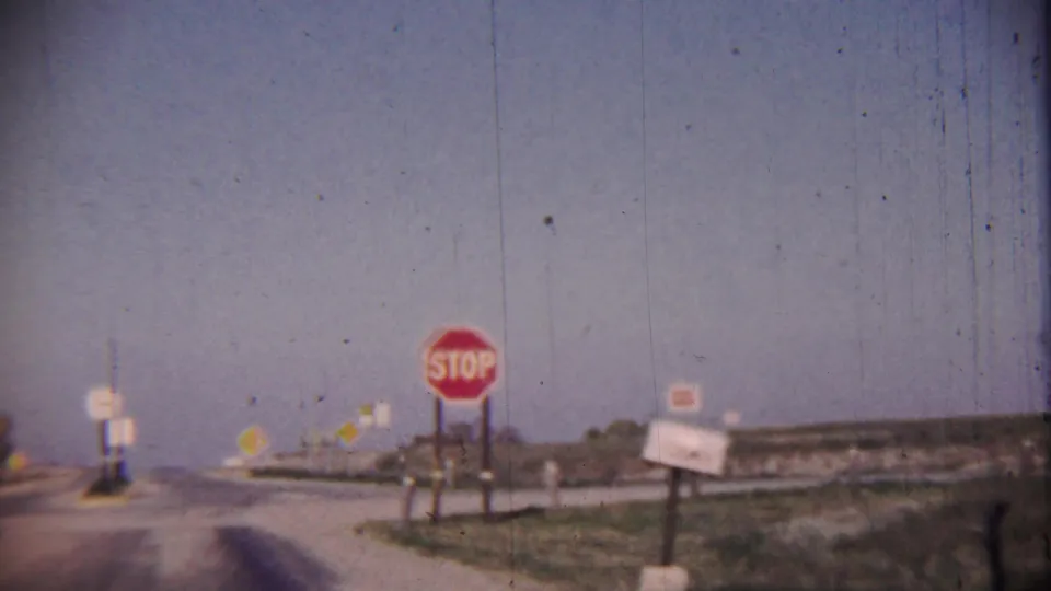 1959 8mm view of a rural Cheyenne County, Kansas intersection with a red stop sign, roadway, grassy shoulder, and pale sky.