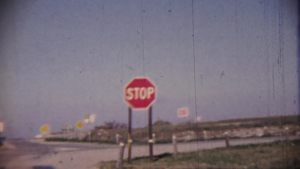 Closer 1959 rural Kansas roadside frame showing a red stop sign, paved junction, grass verge, and distant route markers.