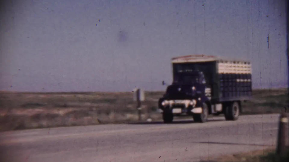 A dark blue truck with a tall cargo body moves along a paved rural highway through flat open Kansas landscape.