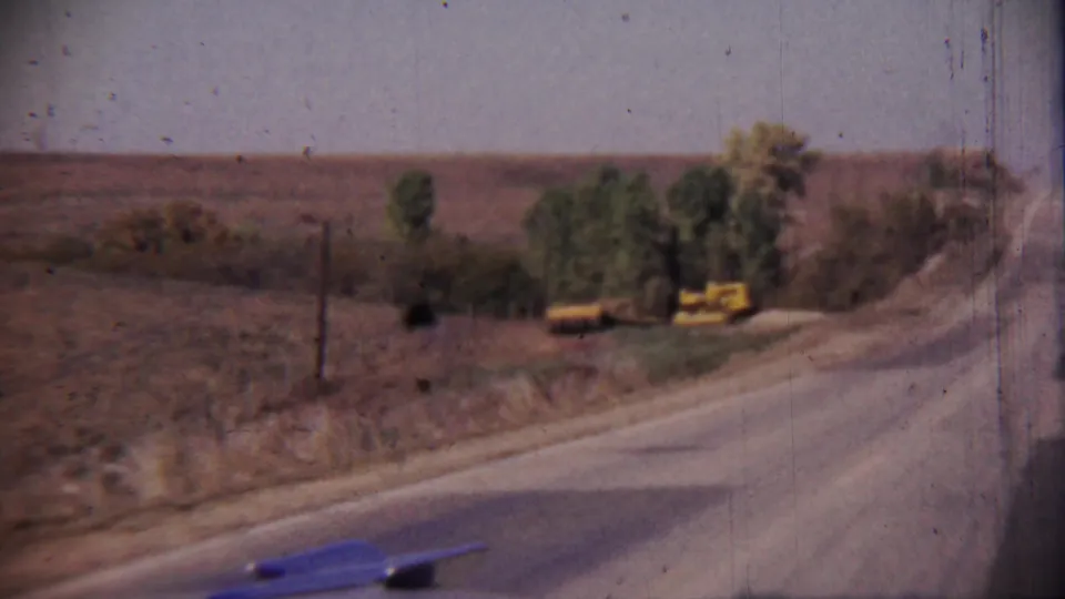 The moving-camera view looks past a rural road shoulder toward open fields, trees, and yellow roadside equipment in Cheyenne County, Kansas.