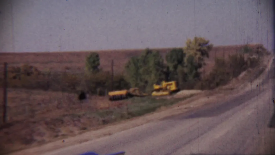 A rural roadside landscape shows a paved road curving along open fields with trees and yellow equipment near the shoulder.