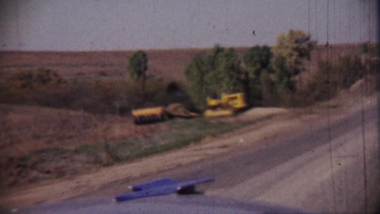 1959 Cheyenne County, Kansas home-movie frame showing a rural road, dry fields, trees, and yellow construction equipment at the roadside.