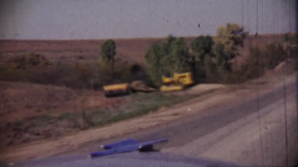 1959 Cheyenne County, Kansas home-movie frame showing a rural road, dry fields, trees, and yellow construction equipment at the roadside.