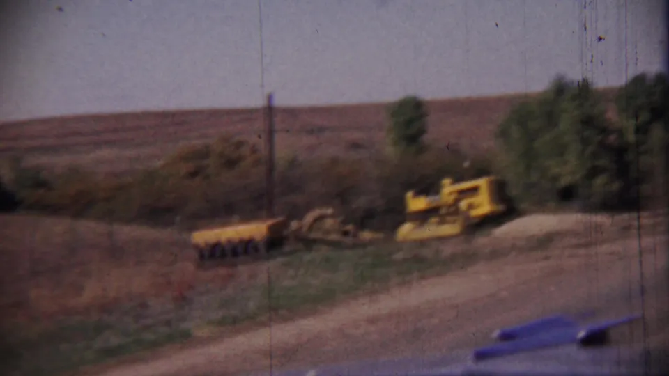 Rural Kansas roadside view with yellow construction machinery, a dirt road edge, scattered trees, and open fields under a hazy sky.