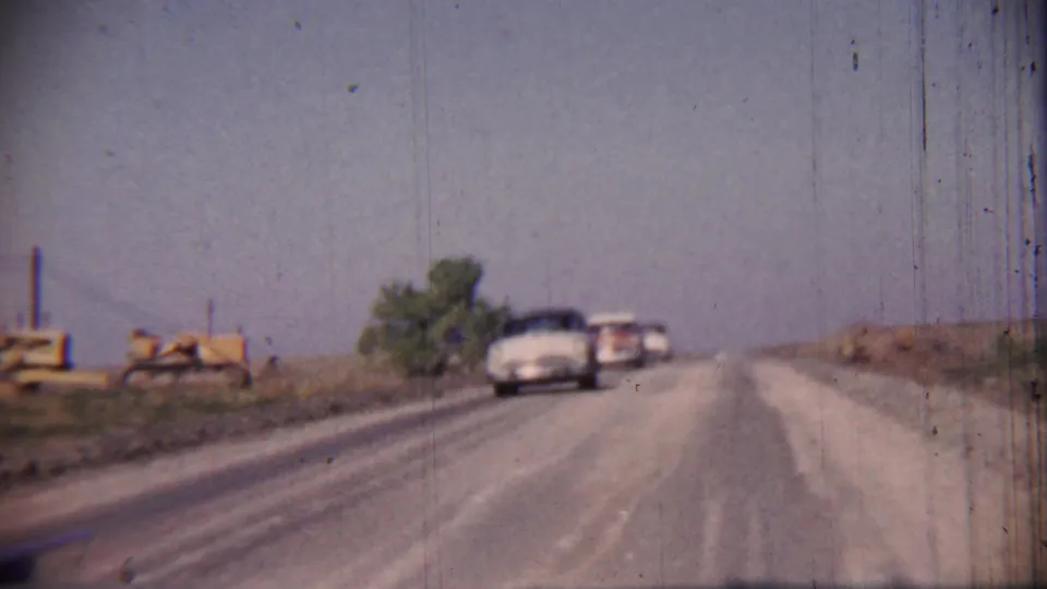1959 rural Kansas road scene with two mid-century cars on a rough unpaved surface and yellow equipment visible along the shoulder.