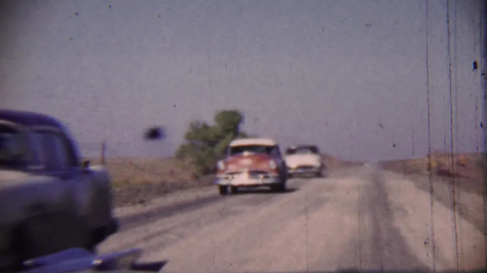 A dark vehicle passes close to the camera while older sedans travel along a rural Cheyenne County road in the 1959 home-movie footage.