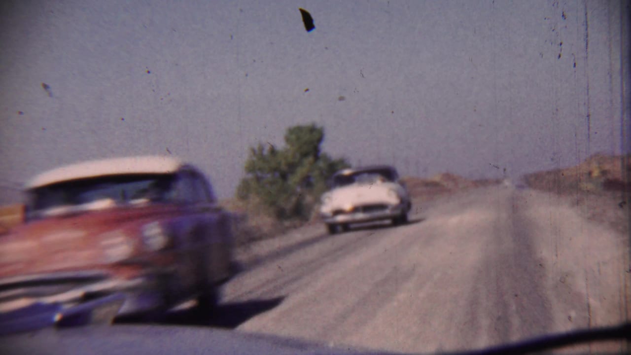Red and white car passes on a rural highway with another car approaching behind it.