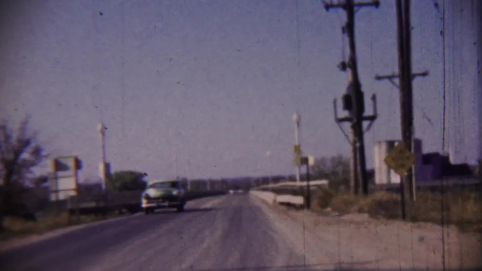 1959 rural Kansas road view with an approaching dark car, roadside signs, utility poles, and a bright open sky.