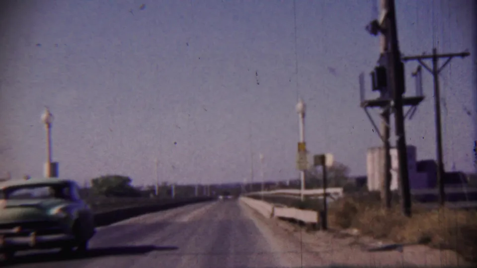 Blue and white mid-century car moves away along a Cheyenne County, Kansas highway with utility poles, small structures, and roadside signs visible.