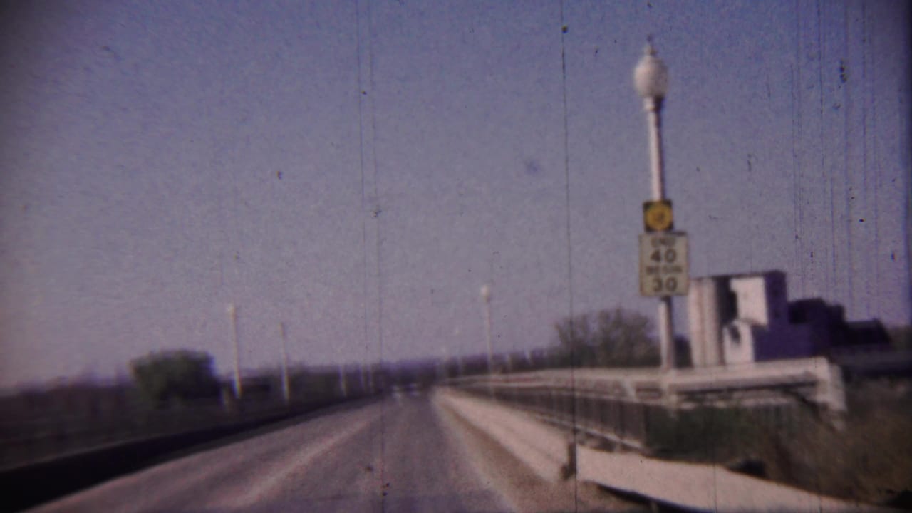 Roadway across a bridge with a speed limit sign, lamp posts, railings, and distant buildings.