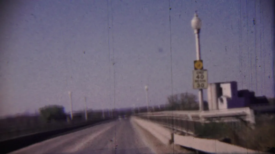 Forward driving view in Cheyenne County, Kansas, with a bridge rail, END 40 BEGIN 30 sign, lamps, and low buildings ahead.