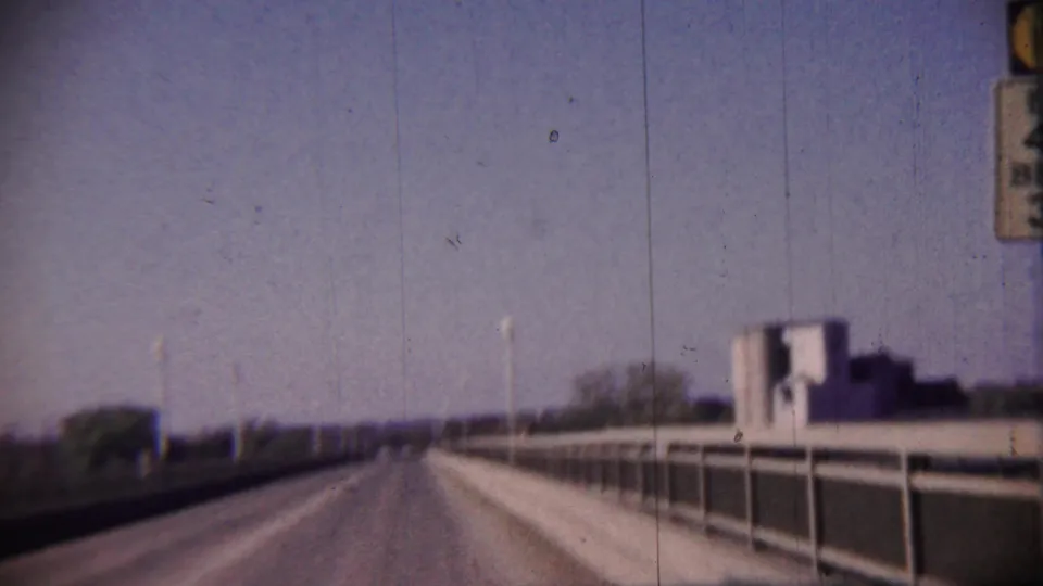 1959 8mm frame from a moving vehicle on a Cheyenne County bridge, showing railings, lamps, trees, and distant structures.