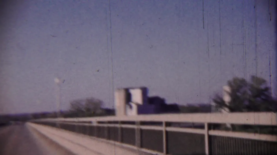 End frame of the Cheyenne County bridge drive, with white guardrail, road edge, a distant low building, tree, and street lamp.