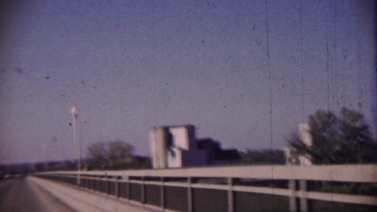 Roadway on a bridge with railing, a light pole, trees, and a distant structure under a pale sky.